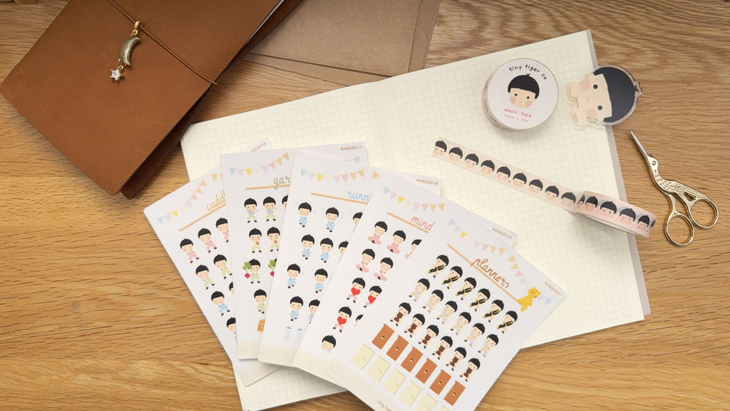 A styled flat-lay of a stationery bundle set featuring five character sticker sheets, an acrylic binder clip, and a washi tape roll, arranged on a light wood desk alongside a brown notebook and gold scissors.